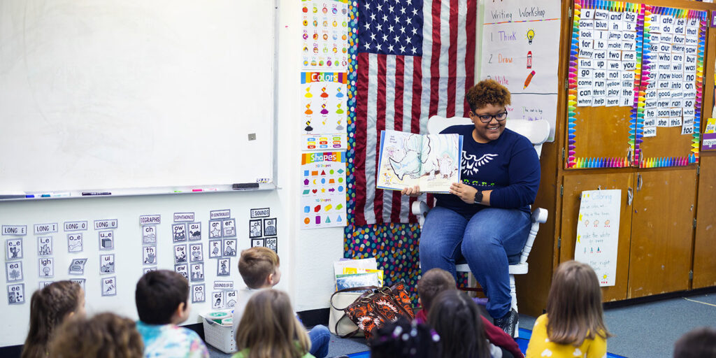 Early elementary teacher reading to class of students sitting on a rug.